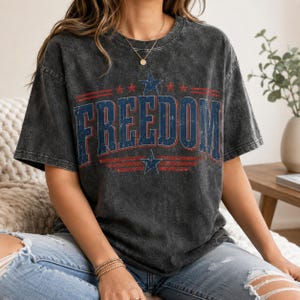 Könnte beinhalten: Ein anthrazitfarbenes T-Shirt in Acid-Wash-Optik mit dem Wort FREEDOM in großen blauen Blockbuchstaben mit roter Umrandung. Das Design enthält rote horizontale Streifen und blaue Sterne über und unter dem Text, was einen patriotischen Grafikdruck ergibt.