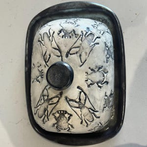 May include: A rectangular ceramic butter dish with a lid. The lid is white with black insect illustrations. The dish sits in a dark grey metal tray. A small, round, black knob is on top of the lid.