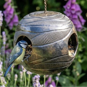 Könnte beinhalten: Ein kugelförmiges Vogelfutterhaus aus dunkler Keramik mit einem strukturierten, blattartigen Muster auf der Oberfläche. Das Futterhaus hat zwei kreisförmige Öffnungen, die mit Vogelfutter gefüllt sind, und ein kleiner blau-gelber Vogel sitzt am Rand.