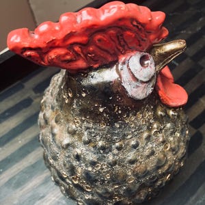 Può includere: Statuetta di gallo in ceramica con corpo bronzo metallizzato strutturato e cresta rosso vivo. L'occhio del gallo è bianco e il becco dorato. La statuetta ha un aspetto rustico e artigianale, adatta all'arredamento della casa.