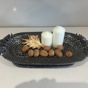 Könnte beinhalten: Eine dekorative schwarze Schale mit strukturierter Oberfläche, die zwei weiße Kerzen, eine Muschel und mehrere Walnüsse enthält. Die Schale hat einen gewellten Rand und schafft eine rustikale und elegante Präsentation.