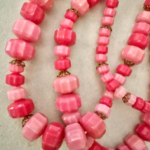 May include: A multi-strand necklace featuring faceted beads in various shades of pink, ranging from light blush to deep magenta. The beads are interspersed with small, intricate gold-tone metal filigree caps between the individual segments.
