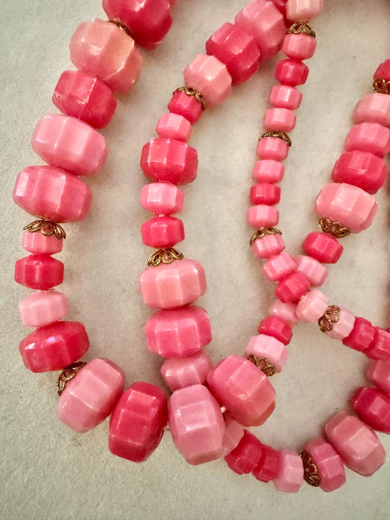 May include: A multi-strand necklace featuring faceted beads in various shades of pink, ranging from light blush to deep magenta. The beads are interspersed with small, intricate gold-tone metal filigree caps between the individual segments.