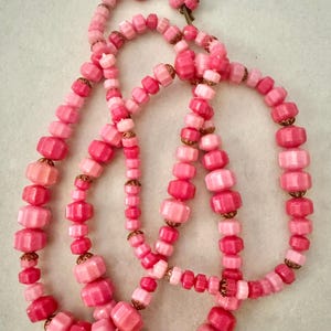 May include: A long necklace featuring a series of faceted beads in varying shades of pink, ranging from light pastel to deep rose. The beads are separated by small, metallic, scalloped-edge spacers and are strung together in a continuous loop.