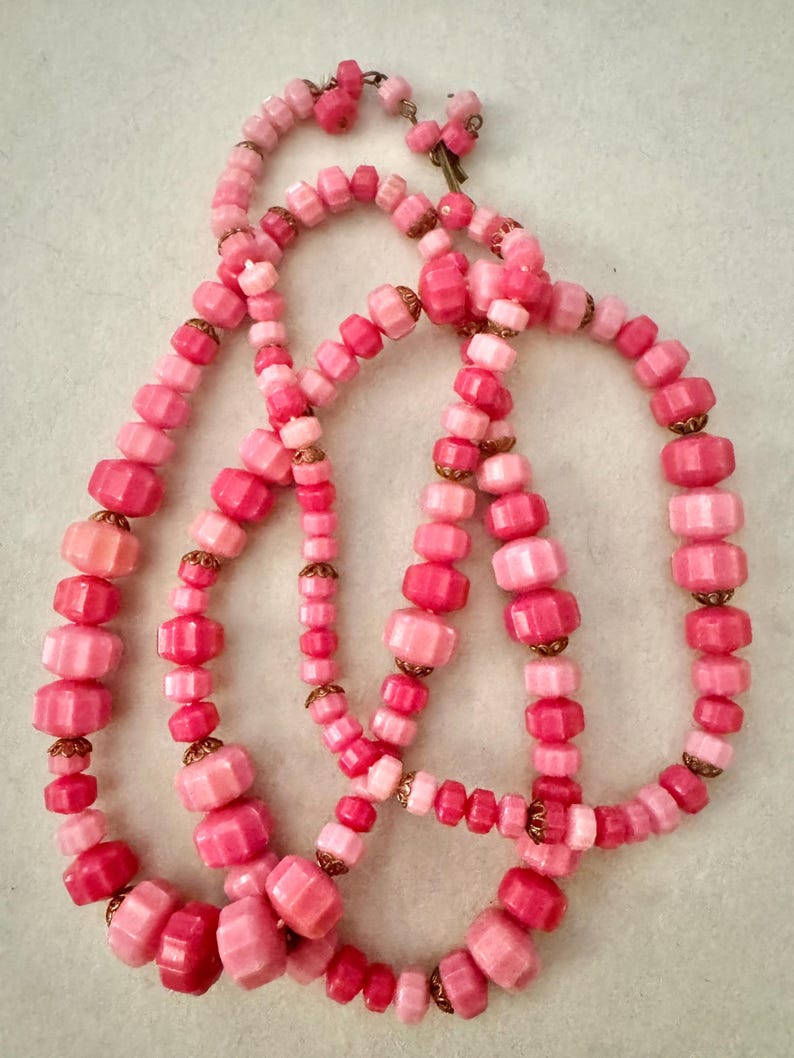 May include: A long necklace featuring a series of faceted beads in varying shades of pink, ranging from light pastel to deep rose. The beads are separated by small, metallic, scalloped-edge spacers and are strung together in a continuous loop.
