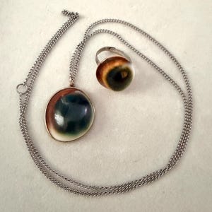 May include: A silver-toned metal necklace with a round pendant and a matching ring. Both pieces feature a smooth, domed inlay with a gradient pattern transitioning from white at the edges to brown and a dark, glossy center resembling an eye.