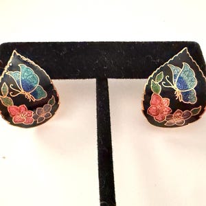 May include: A pair of teardrop-shaped earrings featuring a black enamel background with gold-tone metal outlines. Each earring displays a colorful butterfly in blue and green tones, accompanied by pink and purple floral motifs at the base.