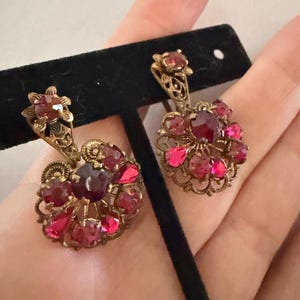 May include: A pair of drop earrings featuring ornate gold-tone metal filigree settings. Each earring is adorned with multiple faceted deep red and pink stones arranged in a circular cluster, with a single matching stone at the top.