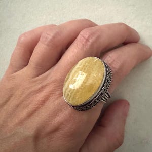 May include: A ring featuring a large, oval-shaped, pale yellow stone with a smooth, polished surface. The stone is set in a decorative, oxidized silver metal band with intricate filigree detailing along the edges and a split shank design.