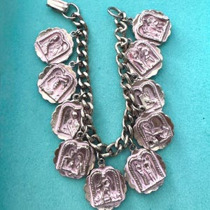 May include: A silver-toned charm bracelet featuring a thick curb chain and nine scalloped, square-shaped charms. Each charm displays a raised, light pink relief depicting various figures and scenes, attached to the chain with small silver jump rings.