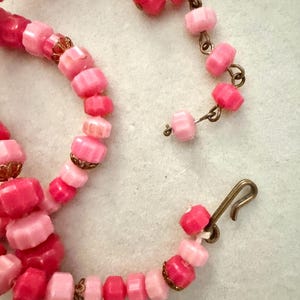 May include: A necklace featuring faceted beads in various shades of pink, ranging from light pastel to deep magenta. The beads are strung together with small, ornate bronze-toned metal caps between some of them, and the piece includes a metal hook clasp.