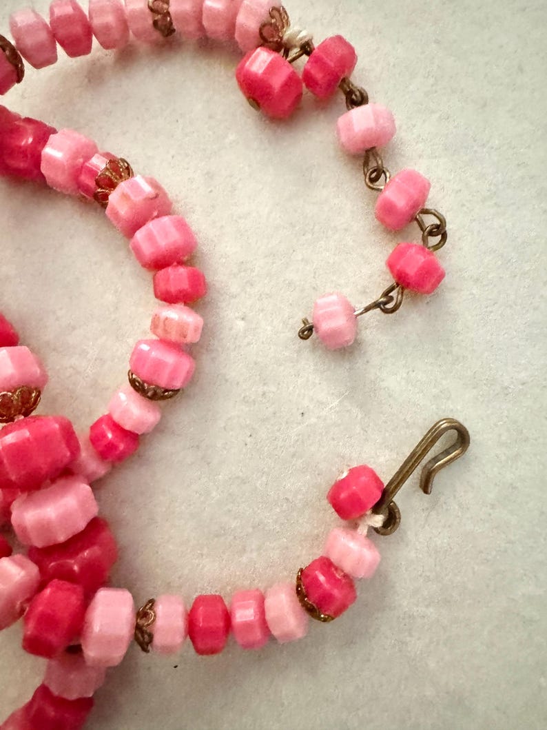 May include: A necklace featuring faceted beads in various shades of pink, ranging from light pastel to deep magenta. The beads are strung together with small, ornate bronze-toned metal caps between some of them, and the piece includes a metal hook clasp.