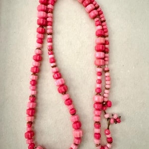 May include: A long necklace featuring a strand of faceted pink beads in varying shades of light and dark pink. The beads are separated by small, metallic gold-toned spacers. The necklace has a simple hook-and-eye clasp closure at the end.