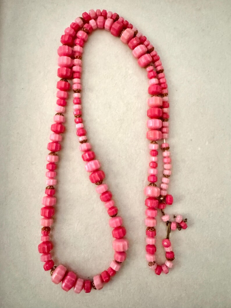 May include: A long necklace featuring a strand of faceted pink beads in varying shades of light and dark pink. The beads are separated by small, metallic gold-toned spacers. The necklace has a simple hook-and-eye clasp closure at the end.