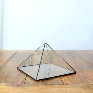 Peut inclure: Un terrarium en forme de pyramide en verre transparent avec des bordures en métal noir. La forme géométrique repose sur une surface en bois. Le terrarium est conçu pour abriter de petites plantes ou des objets décoratifs.