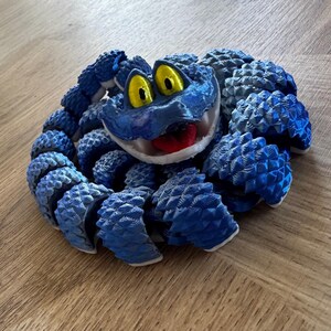 May include: A blue articulated snake toy with yellow eyes and a red tongue. The toy is made of multiple segments, giving it a flexible, realistic appearance. The snake is coiled on a wooden surface.