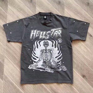 May include: Dark gray t-shirt with the word "HELLSTAR" in a stylized font above a white skeleton graphic. The skeleton is seated in a meditative pose, surrounded by flames. Small white stars are printed on the sleeves.