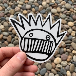 May include: A die-cut vinyl sticker featuring a black and white graphic illustration of a stylized face with a large, toothy grin and a crown-like spiky hairstyle. The design is bold, high-contrast, and features clean, thick black lines.