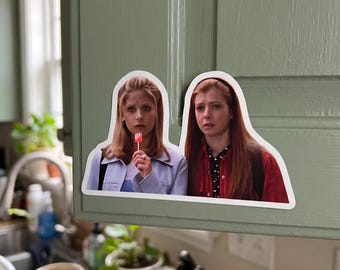 May include: A die-cut vinyl sticker featuring two women with long hair. The woman on the left has blonde hair, wears a light blue jacket, and holds a pink lollipop. The woman on the right has reddish-brown hair and wears a red jacket over a black shirt.