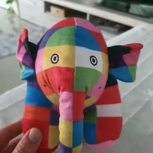 May include: A plush elephant toy featuring a vibrant patchwork design of multicolored squares in red, blue, yellow, green, and pink. The stuffed animal has large ears, a trunk, and embroidered circular eyes with black and white stitching details.