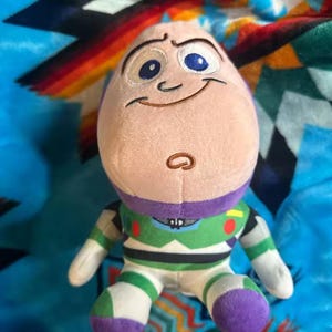 May include: A plush Buzz Lightyear toy, featuring a light purple head, a green and white spacesuit, and purple accents. The toy has embroidered facial features and is set against a colorful patterned blanket.