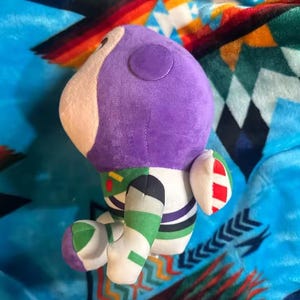 May include: A plush toy of the character Buzz Lightyear from the movie Toy Story. The toy has a purple head, white face, and green, white, and black body. The toy is on a blue blanket with a colorful pattern.