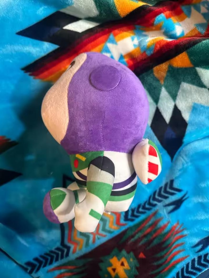 May include: A plush toy of the character Buzz Lightyear from the movie Toy Story. The toy has a purple head, white face, and green, white, and black body. The toy is on a blue blanket with a colorful geometric pattern.
