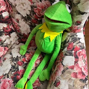 May include: A bright green plush frog toy featuring a yellow felt collar around its neck, large white eyes with black pupils, and long, slender limbs. The frog has a wide, open mouth with a red interior and is constructed from soft fabric.