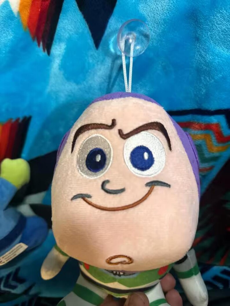 May include: A plush Buzz Lightyear toy with a smiling face, purple accents, and a green and white uniform. The toy has a suction cup attached to the top. The background is a blue blanket with a colorful pattern.