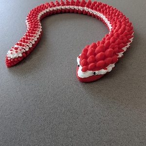 May include: A red and white 3D-printed snake toy. The snake is segmented with a textured pattern, featuring a red body with white accents along the spine and head. The snake's head has small black eyes.