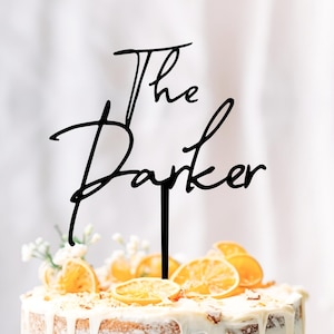 Puede incluir: Un adorno negro para pastel con las palabras "The Parker" en una fuente cursiva. El adorno se coloca sobre un pastel glaseado blanco decorado con rodajas de naranja y pequeñas flores blancas. El pastel tiene un diseño de glaseado goteado.
