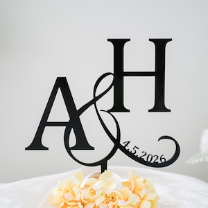 Puede incluir: Adorno para tarta de metal negro con las letras entrelazadas "A" y "H" y un símbolo de ampersand. La fecha "4.5.2026" está inscrita. El adorno se coloca sobre un ramo de flores amarillas, ideal para una boda.