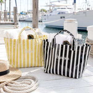 May include: Two rectangular tote bags with vertical stripes, one in yellow and white and the other in black and white. Each bag features two top handles, a white towel inside, and a name embroidered on the front: Reese on the yellow bag and Harper on the black bag.