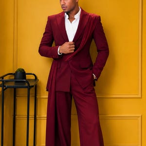 May include: A burgundy suit with a draped accent, paired with a white dress shirt. The suit features wide-leg trousers and is set against a yellow wall. A black metal side table is in the background.