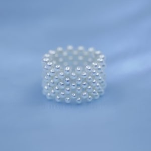 May include: A white beaded ring composed of numerous small, round pearls. The ring is set against a soft, light blue background, creating a contrast that highlights the ring's texture and design. The ring's design is a continuous band of pearls.
