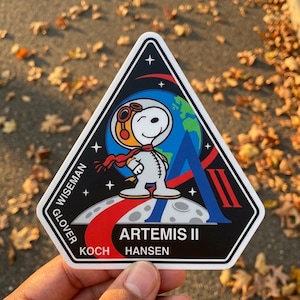 May include: A triangular patch featuring Snoopy in an astronaut suit, with a red scarf and goggles. The patch has the text "ARTEMIS II" and the names "WISEMAN", "GLOVER", "KOCH", and "HANSEN". The background includes a blue Earth and a red and blue "A" shape.