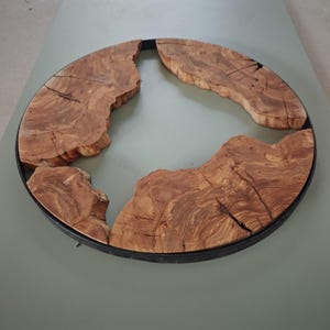 May include: A circular wall art piece featuring a black metal frame enclosing three distinct, irregular segments of natural light brown wood with visible grain patterns, cracks, and raw, uneven edges arranged within the circular boundary.