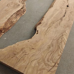 Può includere: Due lastre di legno non rifinite, marrone chiaro, con bordi naturali e venature visibili. Il legno ha un tono chiaro e caldo con striature e nodi più scuri, suggerendo un'estetica rustica o naturale.