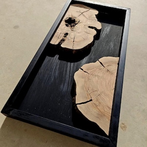Op de afbeelding: Een rechthoekige houten dienblad met een zwart frame. De binnenkant van de lade heeft een donker oppervlak met twee lichtgekleurde, onregelmatig gevormde houten schijven erin. De houten schijven hebben zichtbare nerven en scheuren.