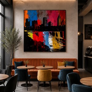 Op de afbeelding: Een vierkant kunstwerk aan de muur met een levendig, abstract silhouet van een stad tegen een kleurrijke, schilderachtige achtergrond van blauwe, rode en gele tinten, boven een lange, oranje gecapitonneerde bank in een modern café.