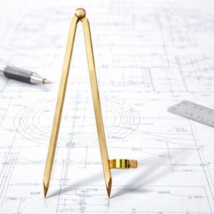 May include: A golden brass compass, used for drawing circles, stands on a blueprint. The compass has two pointed legs connected at the top with a small knob. A pen and ruler are also visible, suggesting drafting or technical drawing.