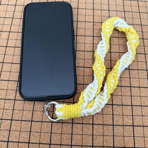 May include: A black smartphone in a case sits beside a yellow and white macrame phone strap. The strap is made of woven cord and has a silver-coloured metal ring.