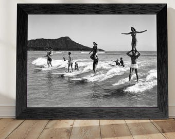 May include: Black and white framed print of people surfing in the ocean. Several surfers are riding waves, some with others on their shoulders. A mountain is visible in the background. The print is in a black frame and is displayed on a wooden floor.