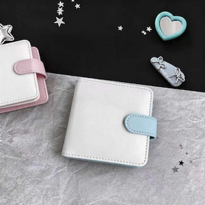 May include: White square-shaped planner with a light blue clasp. The planner is made of a smooth material and has a stitched border. A second planner with a pink clasp is visible in the background. Decorative star and heart-shaped embellishments are scattered around.