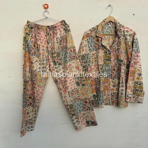 May include: A two-piece pajama set with a floral patchwork design. The set includes a button-down shirt and drawstring pants. The color palette features pink, green, orange, and blue hues. The pajamas are hanging on a wall.