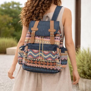 May include: A colorful backpack with a geometric pattern in shades of blue, orange, pink, and brown. The backpack features denim accents, brown leather straps, and side pockets. It has a drawstring closure and buckle details.