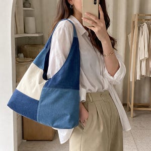 May include: A large shoulder bag with a patchwork design. The bag features panels of light blue, medium blue, and dark blue denim, along with a cream-colored panel. The bag has a single shoulder strap and a spacious interior.