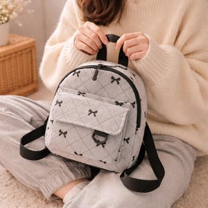 May include: A small, light grey backpack with a quilted pattern and black bow accents. The backpack features a front pocket, black straps, and a top handle. The design is simple and stylish, suitable for everyday use.