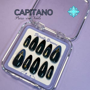 Peut inclure: Un ensemble de dix faux ongles en forme d'amande dans un boîtier en plastique transparent. Les ongles sont bleu marine foncé avec des accents dorés en forme de croix. Le boîtier porte le texte CAPITANO Press-on Nails et Thinails Dreams.