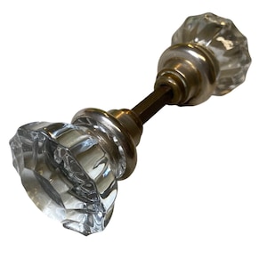 May include: A door knob set featuring two clear, faceted glass knobs with a multi-sided, star-like pattern. The knobs are connected by a central metal spindle and include circular, metallic-toned base plates that sit between the glass and the rod.